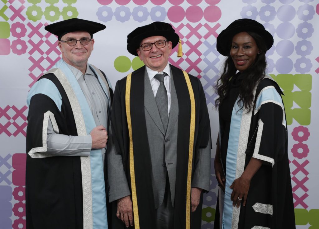 Three people dressed in academic regalia stand smiling against a colourful geometric background. Two men in black hats and capes with pale blue and silver accents flank a woman in a similar outfit. The man in the center wears a suit and a black hat with a gold trim.