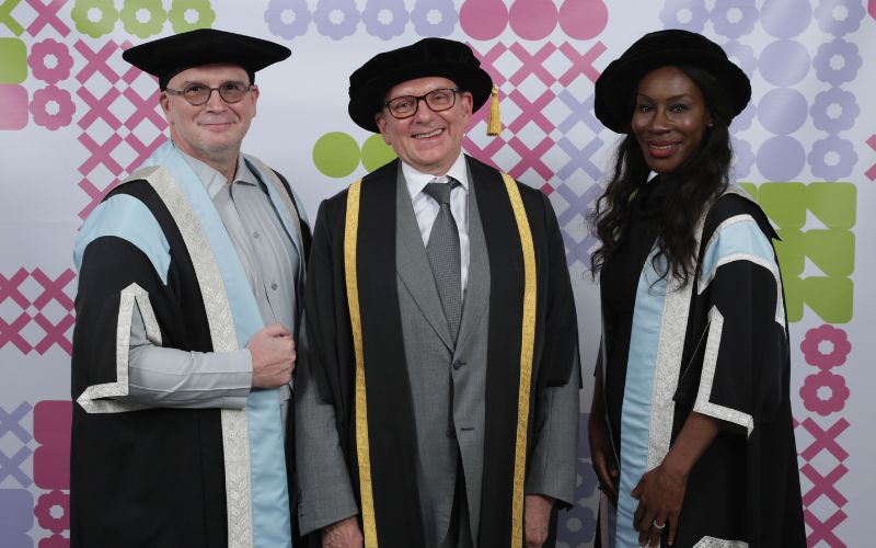 Three people dressed in academic regalia stand smiling against a colourful geometric background. Two men in black hats and capes with pale blue and silver accents flank a woman in a similar outfit. The man in the center wears a suit and a black hat with a gold trim.