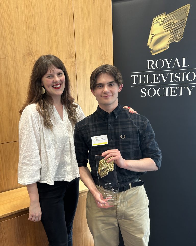 Two people stand in front of a "Royal Television Society" banner. The person on the right, holding a trophy, wears a dark shirt and beige pants. The person on the left, wearing a white blouse and black pants, smiles while standing beside them.