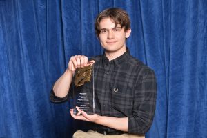 A person with short brown hair and a plaid shirt holds a clear award with gold accents against a blue curtain backdrop. They are smiling slightly and displaying the award prominently.