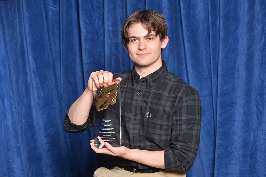 A person with short brown hair and a plaid shirt holds a clear award with gold accents against a blue curtain backdrop. They are smiling slightly and displaying the award prominently.