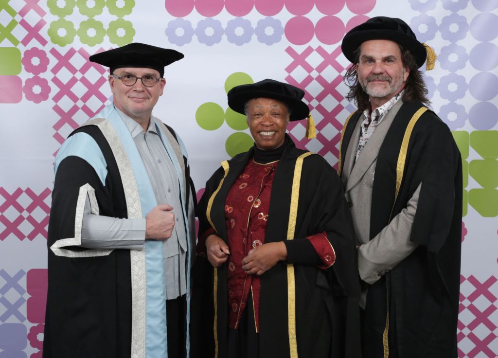 Three people are posing in front of a colourful, patterned background. All are wearing ceremonial academic robes and caps. The person in the center is smiling, while the other two stand on either side looking towards the camera.