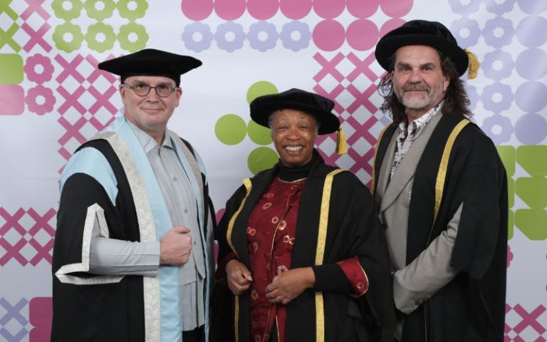 Three individuals dressed in academic regalia pose together in front of a colourful geometric background. They wear black gowns and square academic caps, and all are smiling warmly at the camera. The background consists of patterns in green, pink, and light purple.