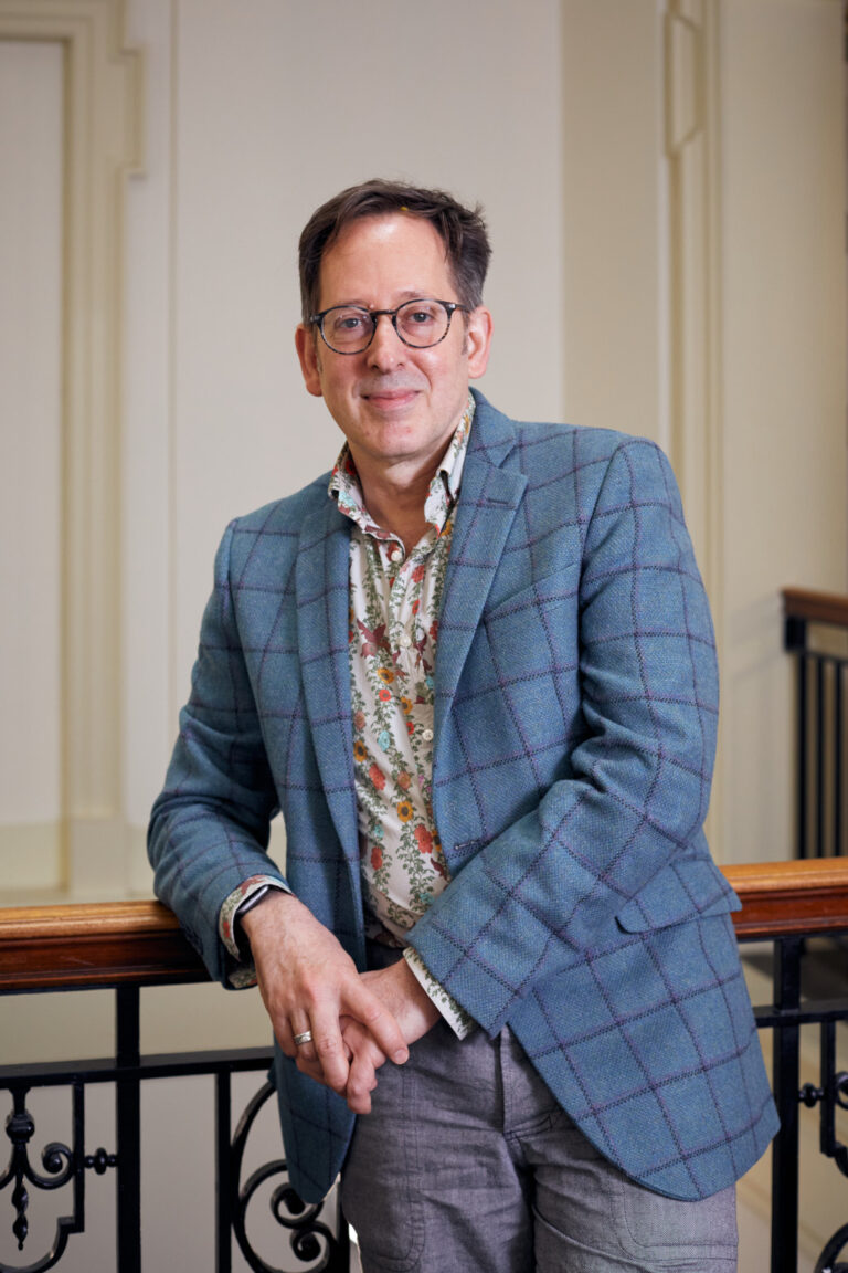 A person with short hair and glasses stands casually leaning against a railing indoors. They are wearing a blue checked blazer over a floral shirt, and light-colored trousers. The setting features neutral-colored walls and a banister.