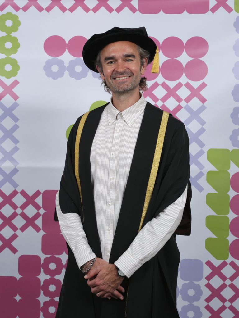 A person with gray hair and a beard is smiling and wearing an academic gown and hat with gold trim. The background features a colourful pattern with pink, purple, and green geometric shapes.