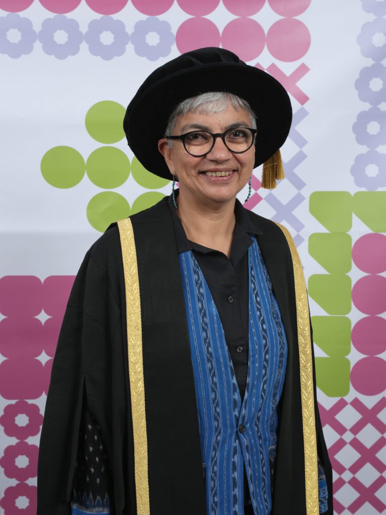 A person stands smiling, wearing glasses and dressed in a black graduation cap and gown with gold trim over a blue and black patterned shirt. The background features a colourful geometric design with circles and squares in pink, green, and purple.