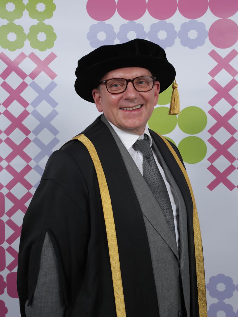 A person wearing academic regalia including a black cap with a gold tassel and a black robe with gold accents smiles while posing against a backdrop with colourful geometric patterns.