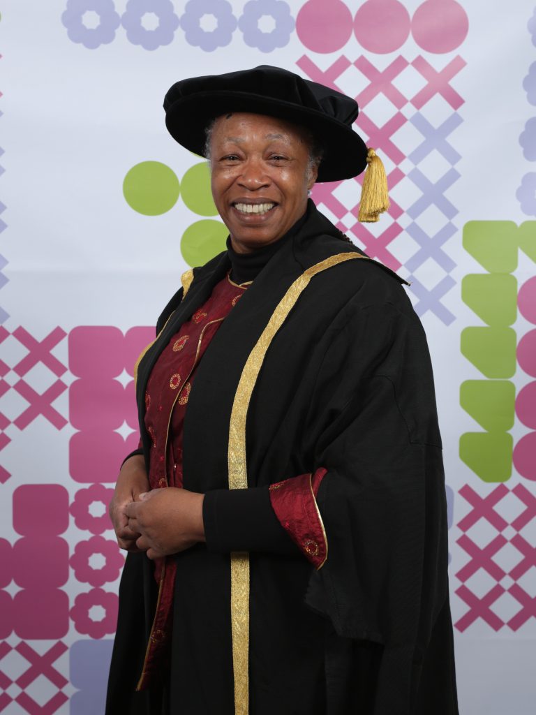 A person in academic regalia is smiling at the camera. They are wearing a black gown with gold trim and a hat adorned with a gold tassel. The backdrop features a colourful pattern of circles and crosses in pink, green, and purple tones.