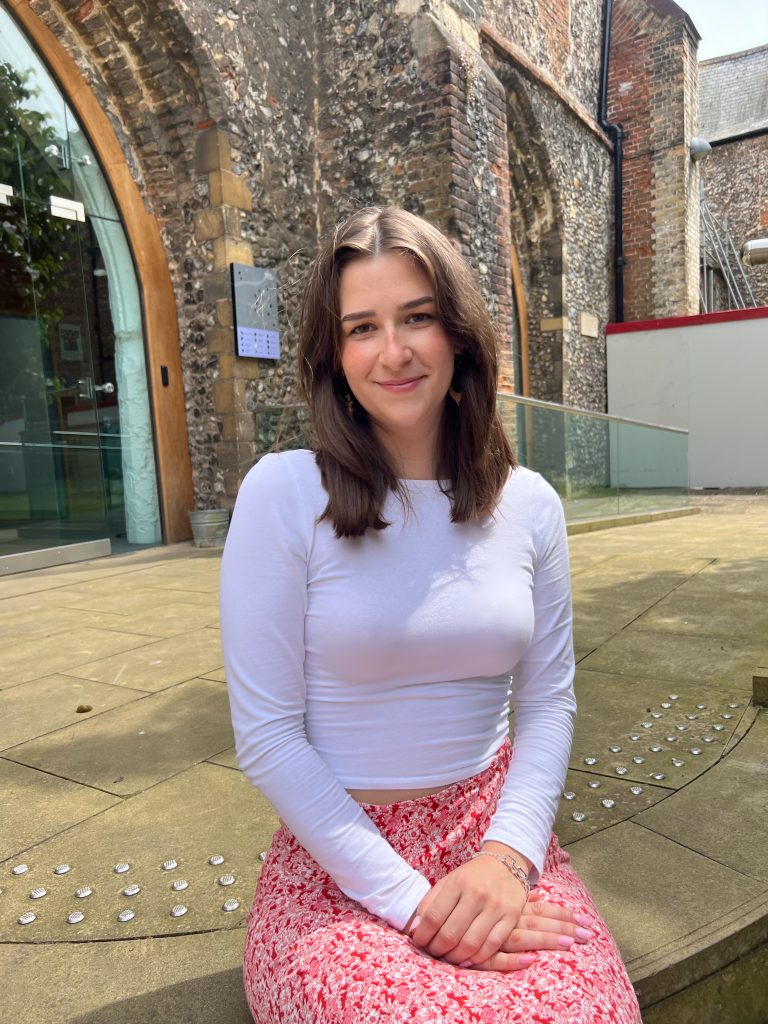 A woman with shoulder-length brown hair and a white long-sleeve shirt sits on stone steps outside a brick building with a glass door. She is wearing a pink and white floral skirt and smiles softly, with her hands resting on her lap.