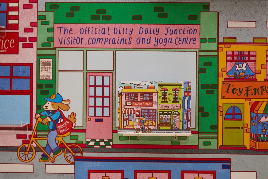 A colourful, cartoonish street scene features a storefront labelled "The Official Dilly Dally Junction Visitor, Complaints, and Yoga Centre." A dog wearing a blue cap and carrying a mail bag rides a bicycle past shops.