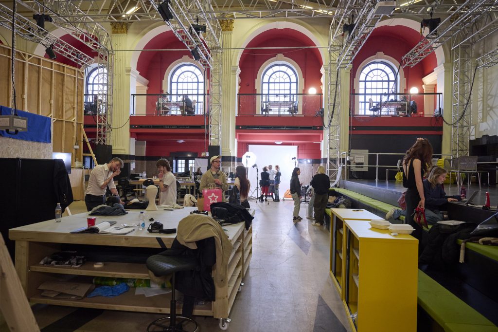 A large room with high ceilings and arched windows is bustling with activity. People are working at various stations, including tables with equipment and a stage area. The space is filled with lighting rigs and scaffolding, suggesting a production or workshop setting.