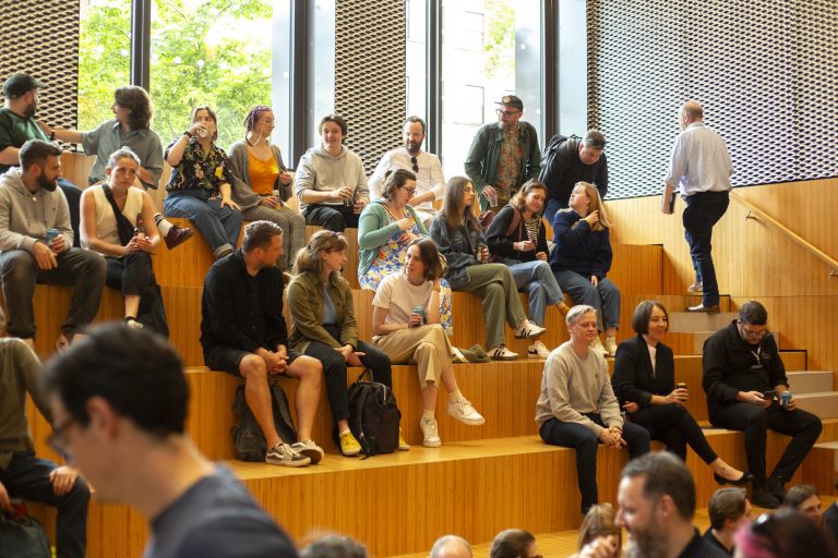 A group of people sits on wooden steps in a large hall with plants visible through the windows. They appear engaged, some holding drinks.