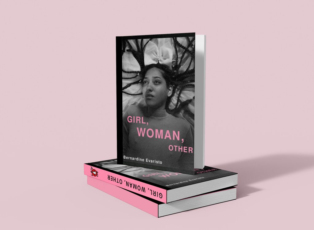 The image showcases Bernardine Evaristo's book "Girl, Woman, Other" standing upright on a pink surface, with a copy lying flat beneath it. The cover features a black and white photograph of a woman with braids looking upward, with the title in pink capital letters.