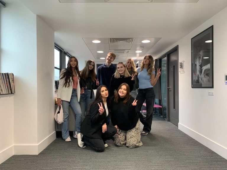 A group of nine people pose together in a hallway, some smiling and making peace signs with their fingers. The hallway has white walls and grey carpet, with some framed pictures hanging on the walls. The group appears to be enjoying a casual moment.