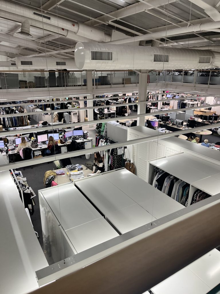 A spacious and busy open-plan office with rows of desks equipped with computers. Employees are seated at their desks working, and the office is surrounded by racks filled with clothing. The setting hints at a fashion or retail workspace.