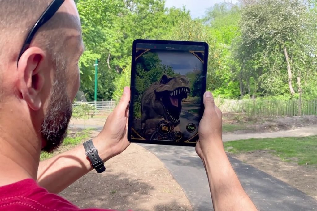 A person holding a tablet outdoors, displaying an augmented reality T-rex dinosaur against a background of greenery and trees. The tablet's screen shows a detailed, roaring dinosaur as if it were in the real world, blending digital and real-life environments.