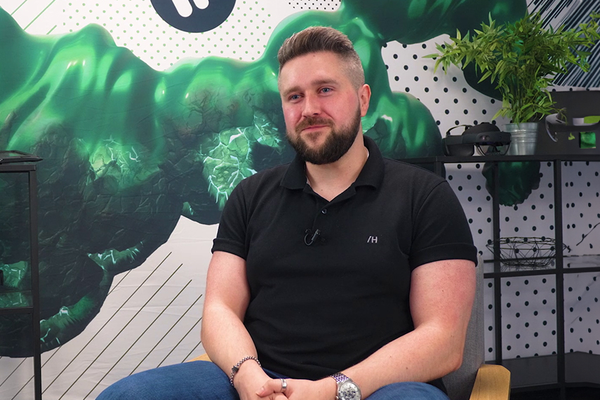 A person with a beard sits on a chair, wearing a black polo shirt and blue jeans. The background features a green and white abstract pattern. There is a shelf and a potted plant to their left.