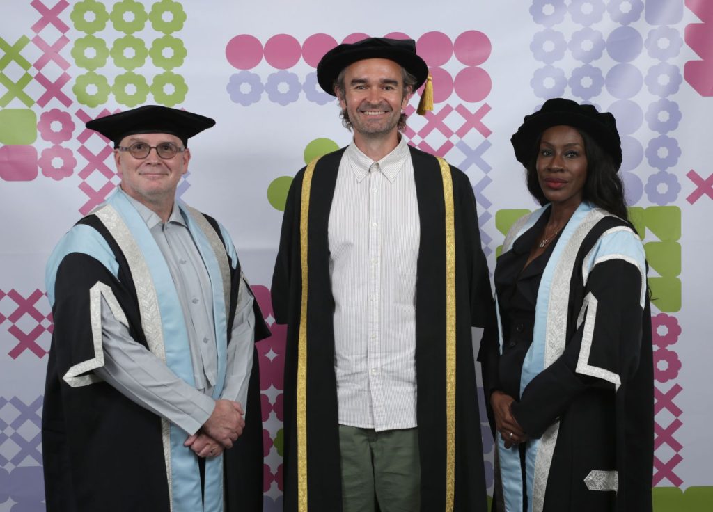 Three people are standing side by side in front of a patterned backdrop. The two on the left and right are dressed in ceremonial academic gowns and hats, while the person in the center is wearing a different academic robe and a wide-brimmed hat. They are all smiling.