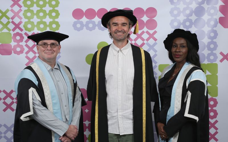 Three people are posing together in academic regalia against a backdrop with geometric patterns. Two people on the ends are wearing light blue and black robes with black hats, while the person in the center is wearing a black and gold robe with a black hat.