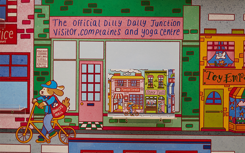 A colourful, cartoonish street scene features a storefront labelled "The Official Dilly Dally Junction Visitor, Complaints, and Yoga Centre." A dog wearing a blue cap and carrying a mail bag rides a bicycle past shops.