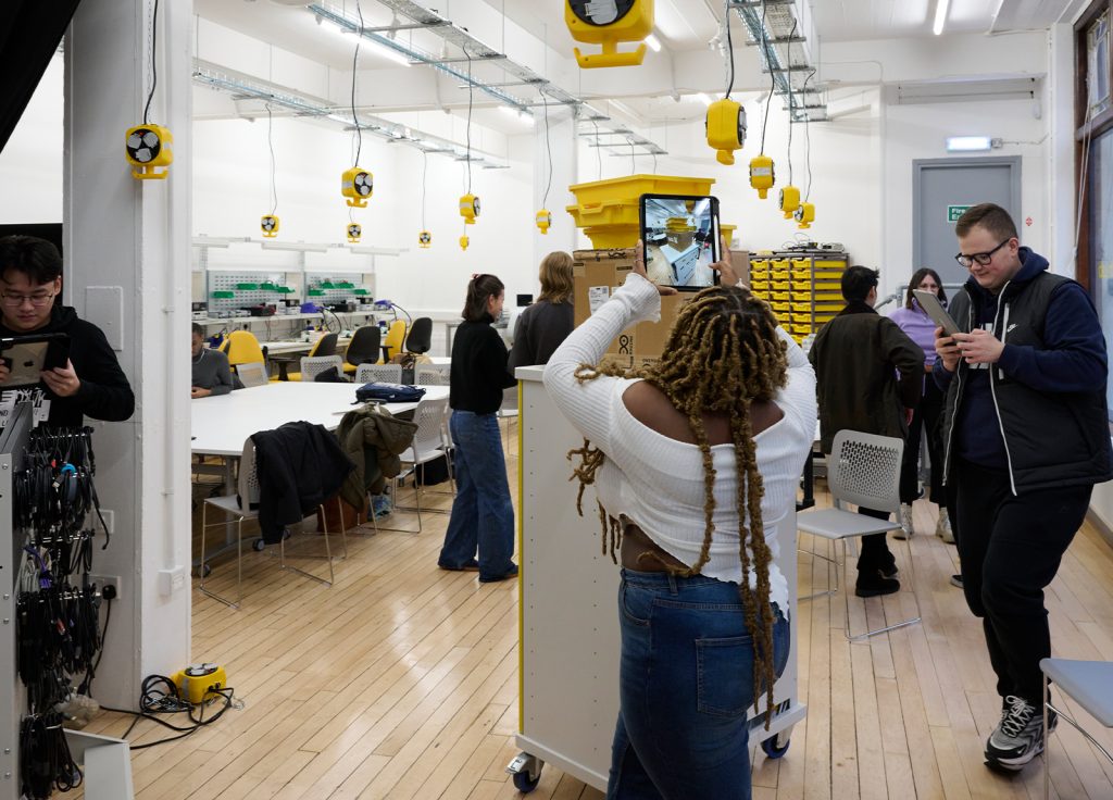 A group of people are working and interacting in a modern, brightly lit classroom or workshop space. There are desks and chairs, various pieces of equipment, and yellow storage bins in the background. In the clearing at the front, one person is taking a photo with a tablet.