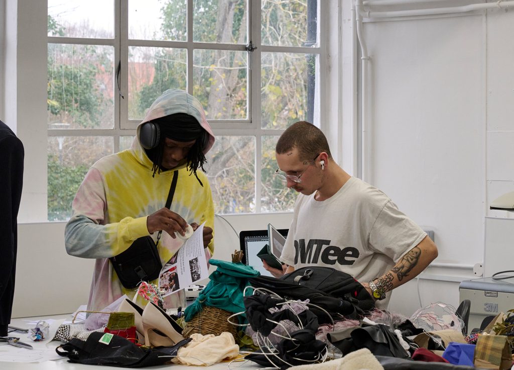 Two individuals are working at a cluttered table covered with fabric, clothing, and accessories. One person wears a tie-dye hoodie and headphones, examining a piece of clothing, while the other in a white t-shirt looks at a document. A window is visible in the background, hinting at the need for some clearing.