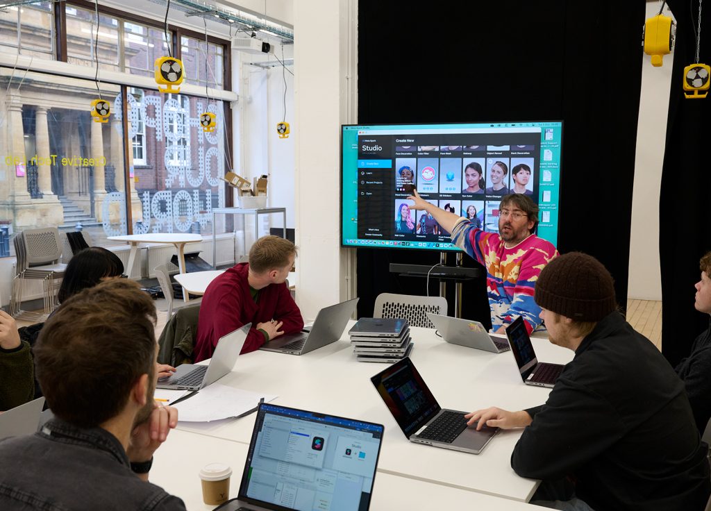 A group of people sits around a white table in a classroom setting, attentively listening to a speaker who points at a large screen displaying a software interface. The room, bathed in natural light from large windows, has an open clearing feel with several laptops open on the table.