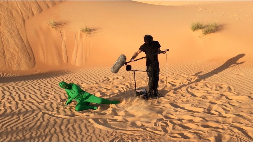 A person in a green bodysuit is lying on the sand in a desert, imitating a fall. Another person dressed in black is standing nearby, holding a boom microphone and wearing headphones. The scene is set against a backdrop of sand dunes with a few sparse tufts of grass.