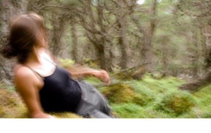 A blurry image depicting a person in a black tank top and greenish pants lying on the ground in a forest. The background shows a dense area with trees and greenery, suggesting a natural setting. The person appears to be in motion or out of focus.