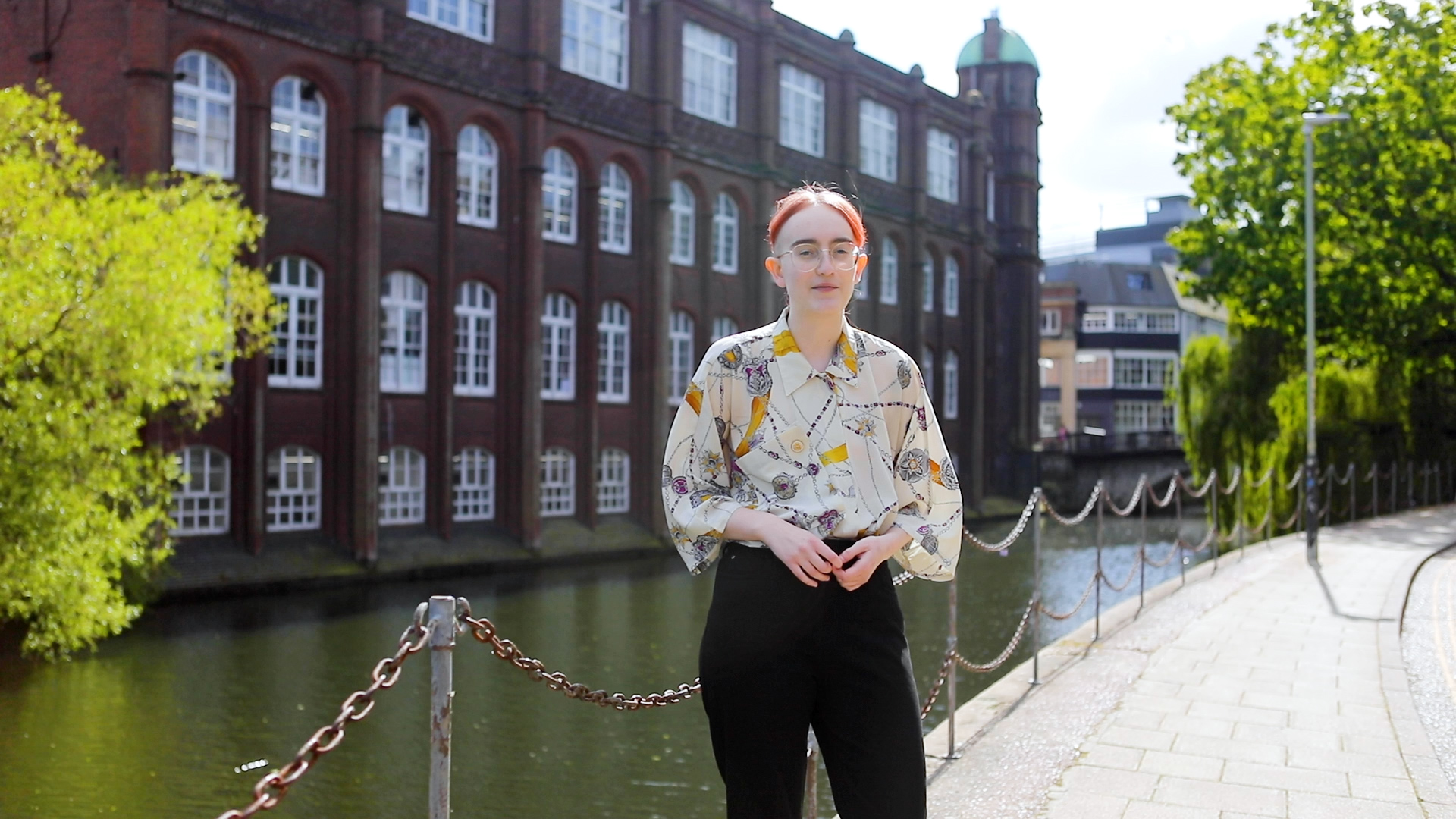 A person with glasses, short hair, and wearing a patterned shirt and black pants stands by a brick building with arched windows along a canal. They are positioned next to a chain-link barrier and a sidewalk, with greenery and water in the background.