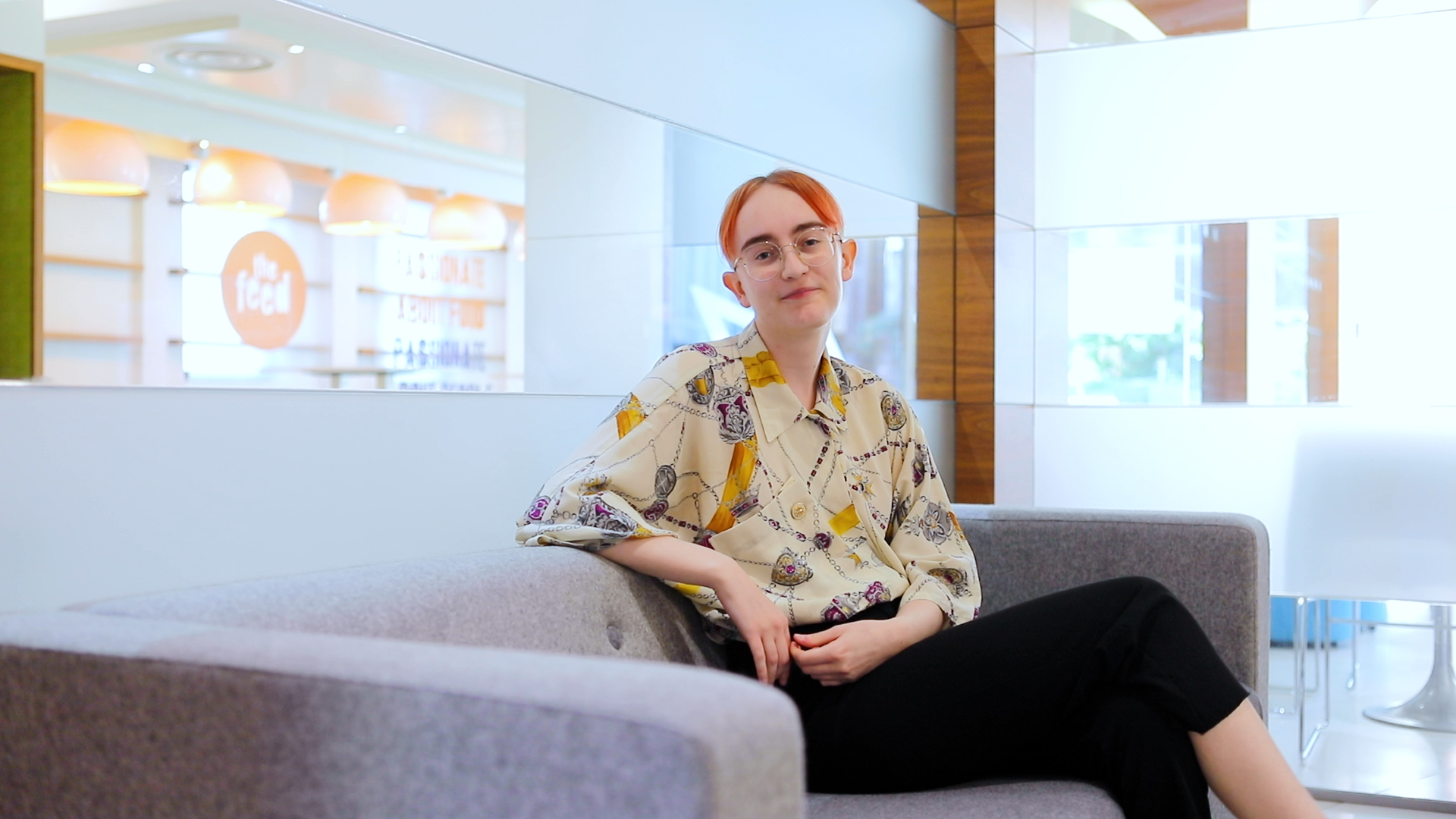 A person with short reddish hair and glasses is seated on a gray couch, smiling. They are wearing a patterned blouse and black pants. The background features a reflective wall and a brightly lit space, possibly a modern lobby or waiting area.