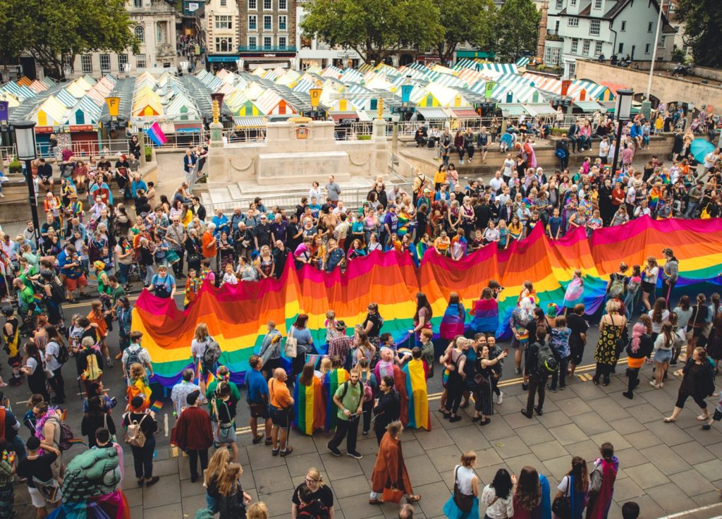 A large crowd of people displays a lengthy rainbow flag during a vibrant outdoor event. Stalls and flags line the background in front of a historic building with colourful decorations, suggesting a festival or parade. The scene is festive and diverse.