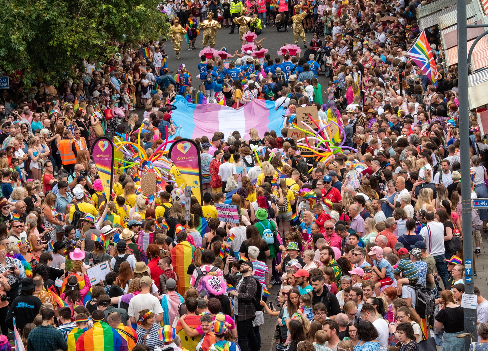 Aerial view of a crowded street during a vibrant pride parade featuring people in colourful outfits, rainbow flags, banners, and a large transgender pride flag. Participants and spectators fill the street with enthusiasm and lively decorations.