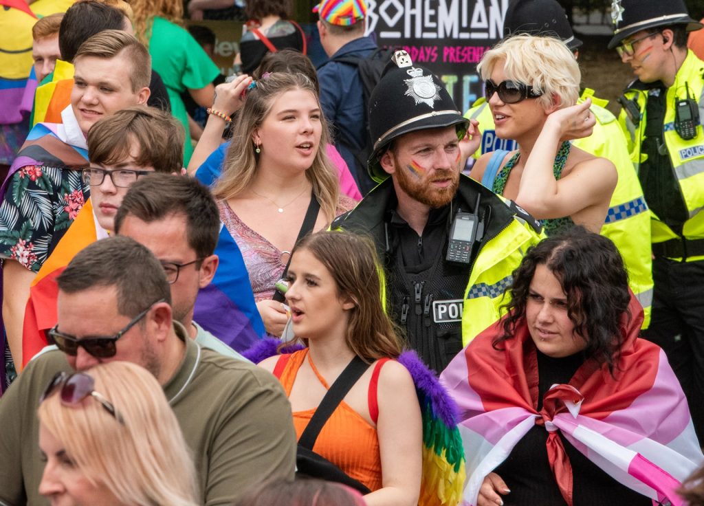 A diverse group of people, many wrapped in rainbow and LGBTQ+ flags, participates in a public event. Several police officers are present. The background includes a "Bohemian" sign and dense crowd. The atmosphere appears festive.