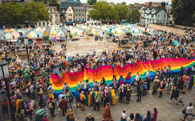 A vibrant outdoor pride event features a large crowd of people gathered around, many holding up an oversized rainbow flag. Colorful market stalls and trees line the background, and attendees are dressed in a variety of colourful outfits, celebrating diversity and inclusion.