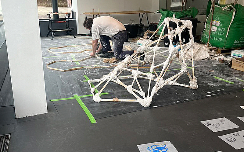 A person kneels on the floor working with wooden frames and white material strands in a room under construction. Green tape marks sections on the dark floor, and various tools and materials are scattered around, including large green bags