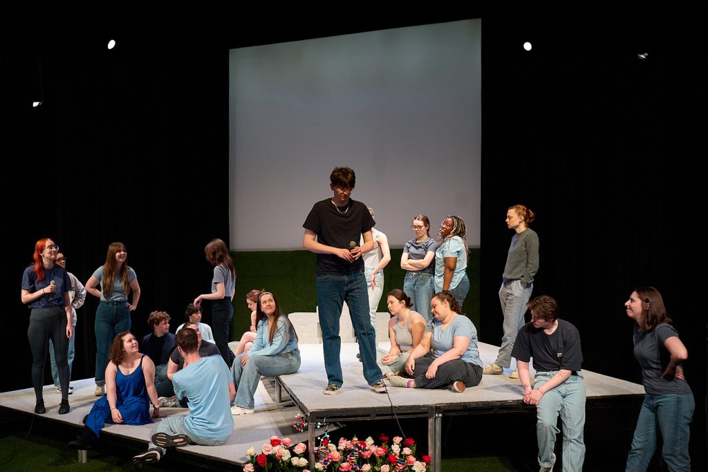 A group of individuals dressed casually are gathered on a raised theatre platform, engaging in various conversations and activities. Some are standing, some are sitting, and a few are interacting with each other.