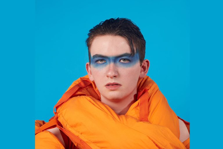 A person with blue face paint across their eyes, wearing an orange jacket, stands against a blue background. Their expression is neutral, and their hair is slicked back. The bold colors create a striking contrast.