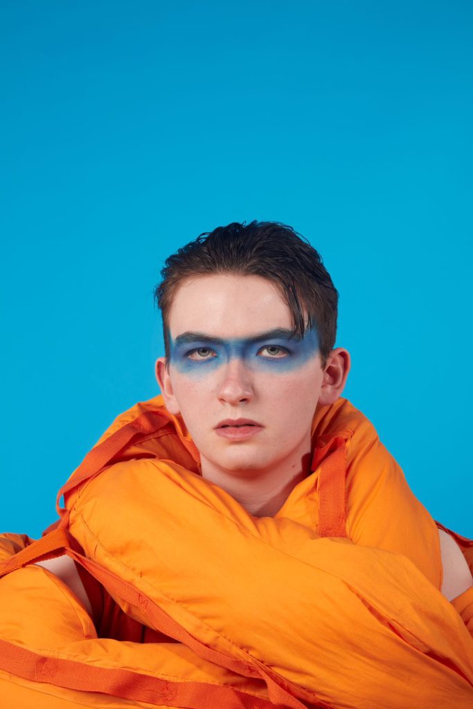 A person with blue face paint across their eyes, wearing an orange jacket, stands against a blue background. Their expression is neutral, and their hair is slicked back. The bold colors create a striking contrast.