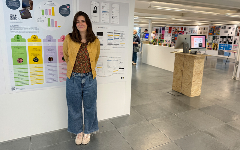 A woman with shoulder-length brown hair, wearing a yellow cardigan, patterned top, and wide-leg blue jeans stands smiling in front of a poster display. The setting appears to be an exhibition space with other posters and a computer on a wooden stand in the background.