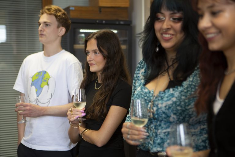 Four people stand closely together in a room, each holding a glass of wine. They appear engaged and attentive.