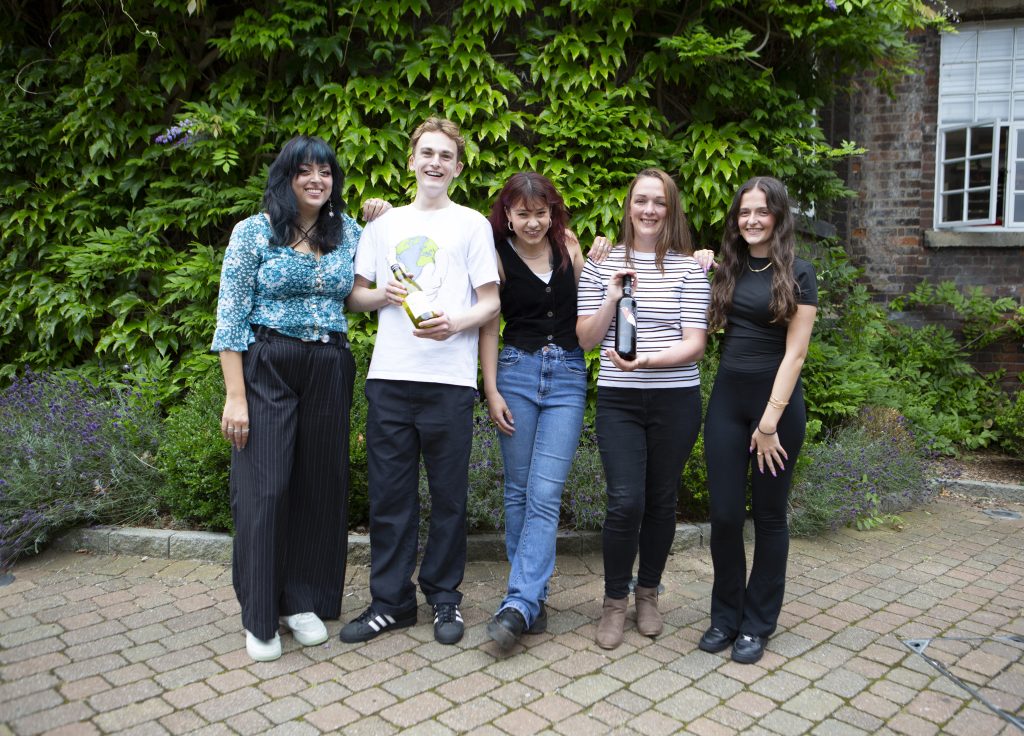 A group of five people stand close together outdoors, smiling at the camera. They stand on a paved area with greenery, including a leafy wall, in the background. They appear cheerful and casually dressed, with some holding wine bottles.