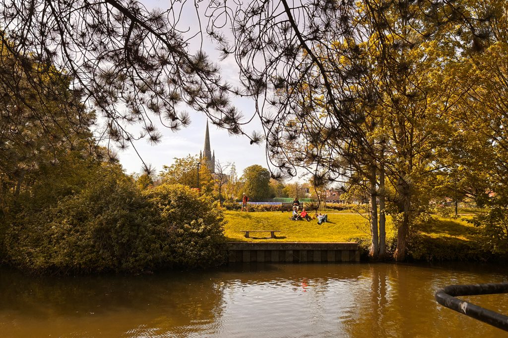 A peaceful park scene with people lounging on a grassy hill near a river. The image features tall, leafy trees framing the edges and a prominent cathedral spire in the background against a light blue sky.