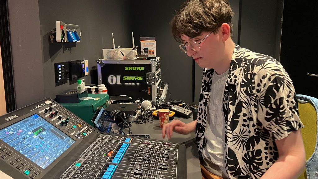 A person wearing a patterned shirt and glasses is adjusting sound levels on a professional audio mixing console in a dimly lit room.