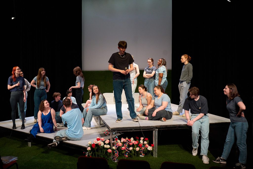 A group of individuals dressed casually are gathered on a raised theatre platform, engaging in various conversations and activities. Some are standing, some are sitting, and a few are interacting with each other.