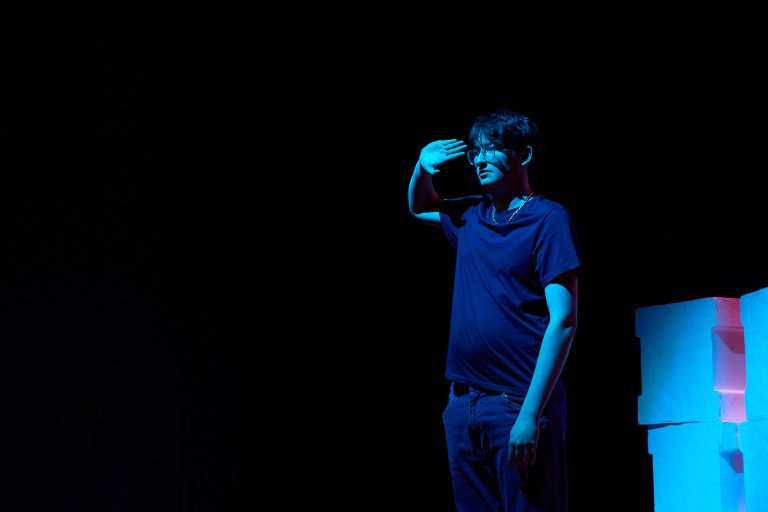 A person stands in a dark space, illuminated by blue lighting. They are saluting with their right hand and wearing a dark-colored t-shirt and jeans.