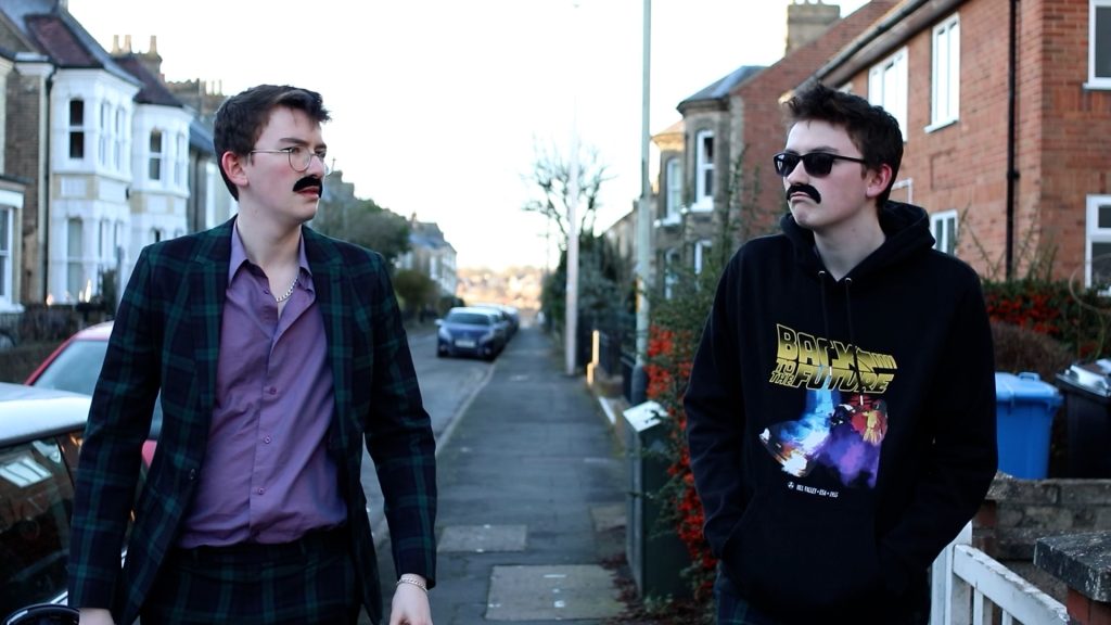 Two young adults with fake mustaches walk down a suburban street. The person on the left wears glasses, a plaid suit, and an open-collared shirt, while the person on the right wears sunglasses, a hoodie with a graphic design, and plaid pants.