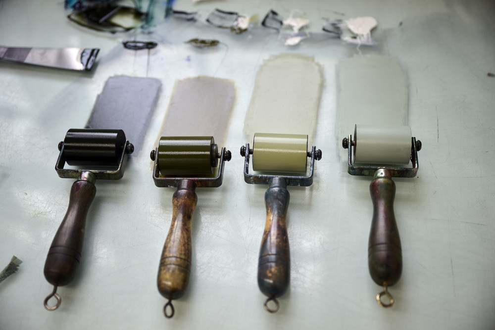 Four ink printing rollers with wooden handles are lined up on a table. Each roller is coated with a different shade of paint, ranging from black to light gray. The paint has been spread beneath each roller. Paint spatulas and a palette are visible in the background.