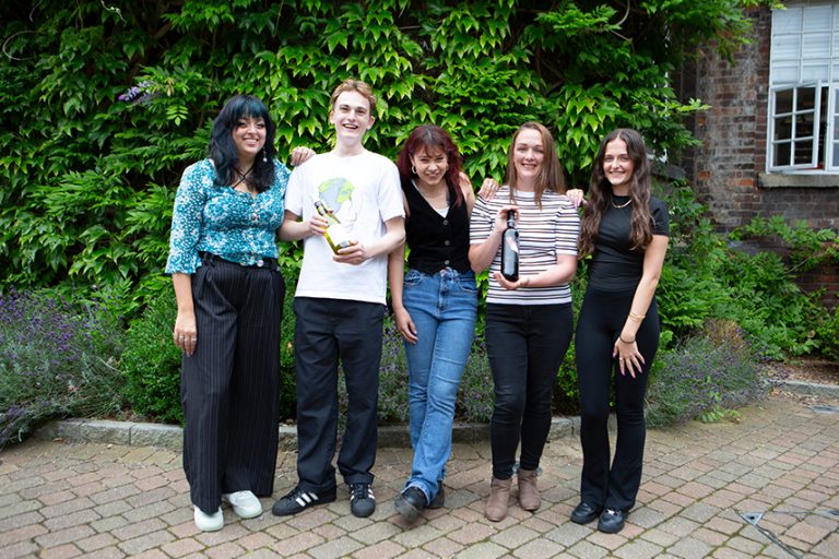 A group of five people stand close together outdoors, smiling at the camera. They stand on a paved area with greenery, including a leafy wall, in the background. They appear cheerful and casually dressed, with some holding wine bottles.