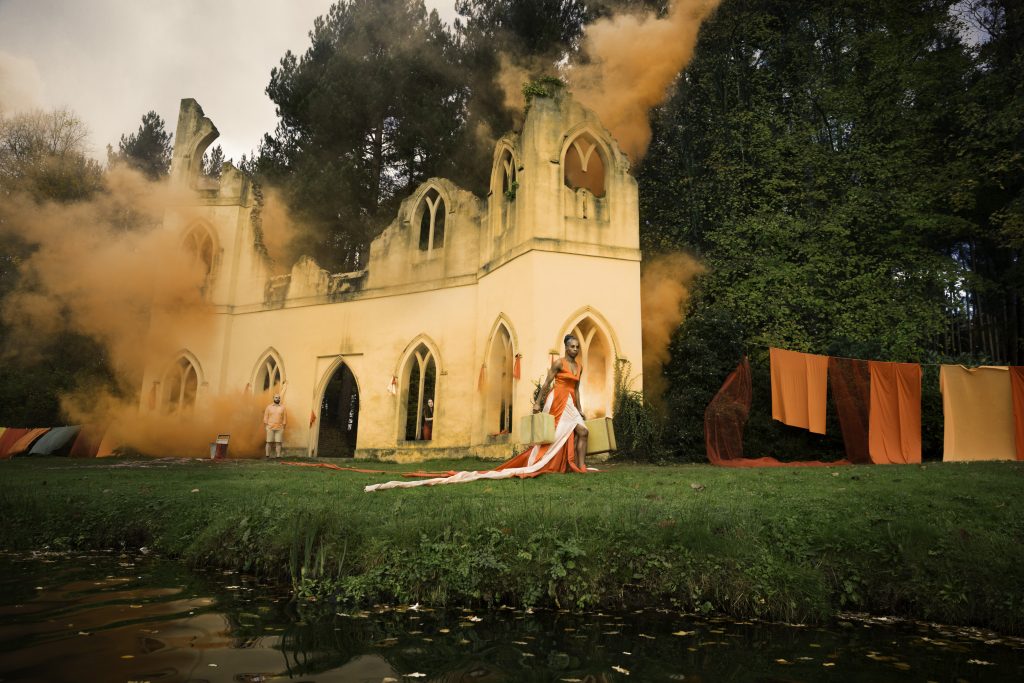 An old, abandoned building with partially ruined walls surrounded by trees has orange smoke billowing from its windows. In front of the building stands a person dressed in orange and white garments with a long trail. Colored fabrics hang nearby.